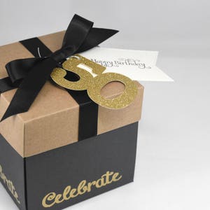 May include: A two-tone gift box with a black base and a tan lid, tied with a black ribbon. A gold glitter "50" and a tag that says "Happy Birthday" are attached. The word "Celebrate" is printed on the box.