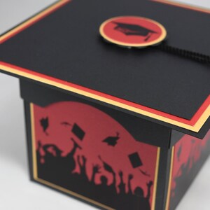 May include: A black graduation cap-shaped gift box with a red and gold trim. The box features a red silhouette design of graduates and mortarboards. A black tassel hangs from the cap's center.