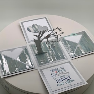 May include: A handmade wedding explosion box with a tree and birds design. The box features a gray and silver color scheme, with the names "Brenda & Eric" and the phrase "Here's to love & laughter and happily ever after."