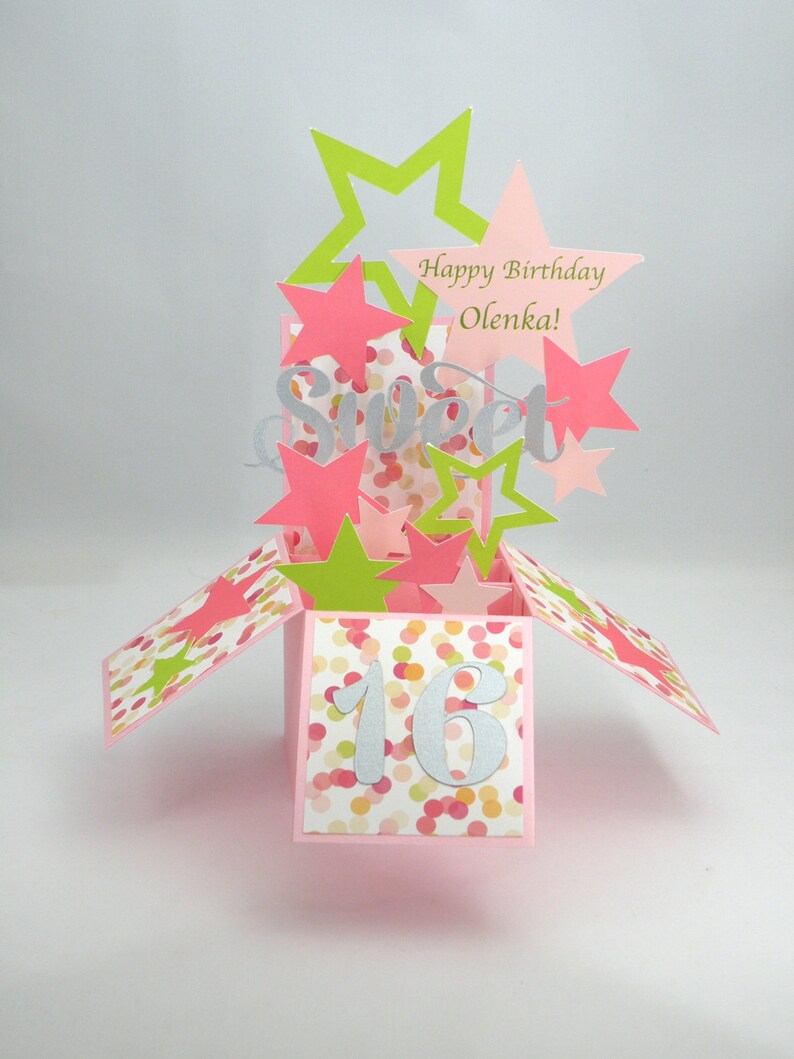 May include: A pink pop-up card with a "Sweet 16" theme. The card features pink, green, and silver star cutouts, with the words "Happy Birthday Olenka!" and "Sweet" displayed. The card is decorated with colorful confetti.