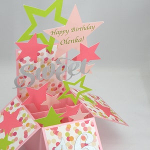 May include: A pop-up birthday card with pink, green, and silver accents. The card features star cutouts, the word "Sweet", and the text "Happy Birthday Olenka!" The card is decorated with a confetti pattern and the number 16.