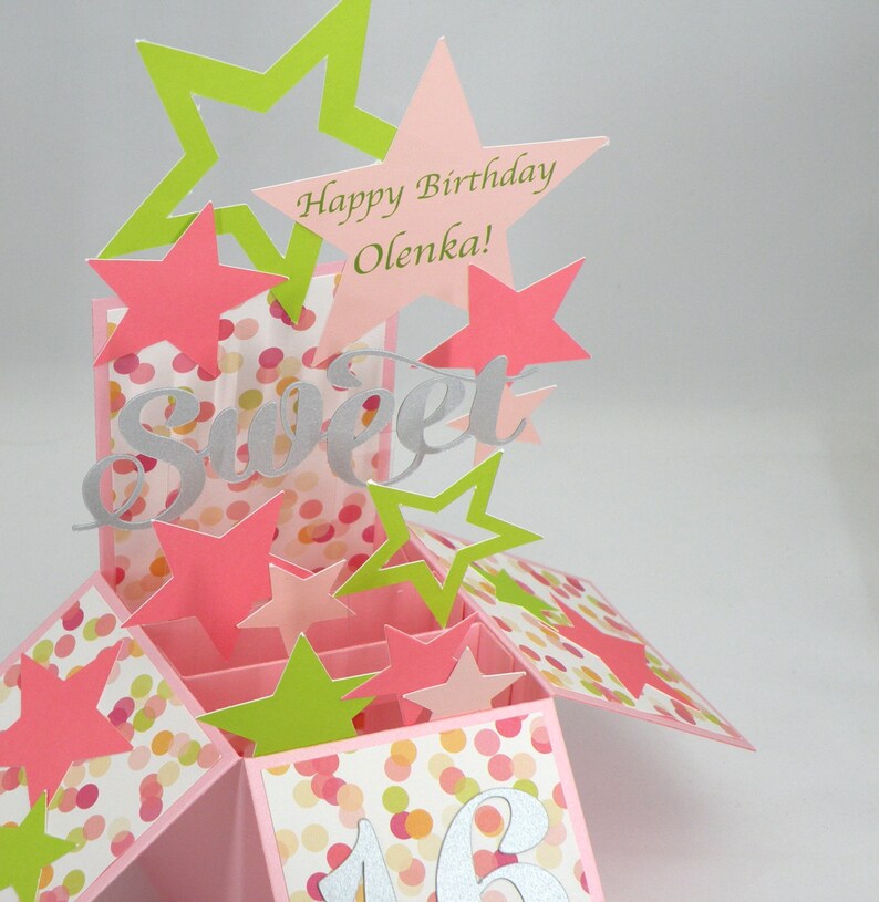 May include: A pop-up birthday card with pink, green, and silver accents. The card features star cutouts, the word "Sweet", and the text "Happy Birthday Olenka!" The card is decorated with a confetti pattern and the number 16.