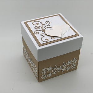 May include: A square gift box with a white lid and a tan base. The lid features a tan square with a white swirl design and two white hearts. The base is decorated with a white floral design.