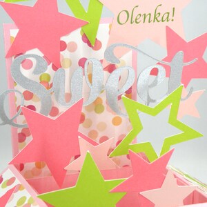 May include: A handmade pop-up card with a "Sweet" silver script design and the name "Olenka!" in green. The card features pink, green, and white star cutouts and a polka dot background. The card is a festive and celebratory design.