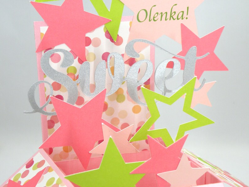 May include: A handmade pop-up card with a "Sweet" silver script design and the name "Olenka!" in green. The card features pink, green, and white star cutouts and a polka dot background. The card is a festive and celebratory design.