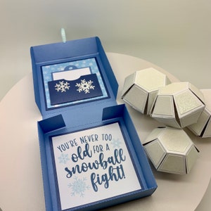 May include: A blue, open gift box with a card that reads "You're never too old for a snowball fight!" and snowflake accents. Several white, geometric ornaments are to the right. The box has a small pocket with snowflake cutouts.