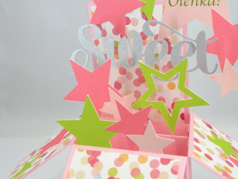 May include: A pop-up card with a pink and white polka dot pattern, featuring the word "Sweet" in silver glitter. The card is decorated with pink and green star cutouts, creating a festive and celebratory design.