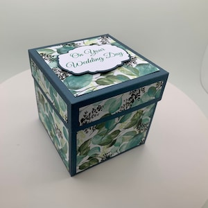 May include: A square gift box with a dark teal frame and a lid. The box is decorated with a pattern of green eucalyptus leaves and dark green accents. The lid features a white label with the text "On Your Wedding Day" in green script.