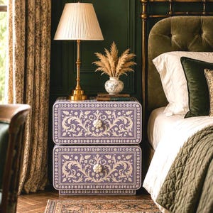 May include: A two-drawer nightstand featuring a light purple finish with intricate white floral inlay patterns, two white flower-shaped knobs, and a rectangular frame. The piece sits beside a bed with a green velvet headboard and bedding.