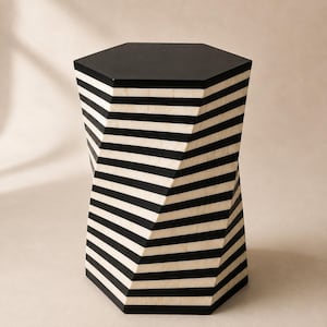 Puede incluir: Una mesa auxiliar hexagonal con una silueta retorcida en forma de reloj de arena. El exterior está decorado con un patrón repetitivo de rayas horizontales negras y color crema. La superficie superior es un hexágono negro plano y sólido.