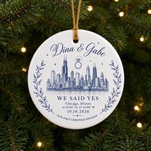 May include: A round white ceramic ornament with a gold hanging cord. The design features a blue Chicago skyline illustration, a diamond ring icon, and the text: Dina & Gabe, WE SAID YES, Chicago, Illinois, 41.8781 N, 87.6298 W, 10.10.2026, OUR FIRST CHRISTMAS ENGAGED.