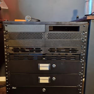 Può includere: Un armadio rack per server nero con diversi pannelli modulari, incluse sezioni ventilate, due cassetti di stoccaggio con serratura e maniglie argentate, e un cassetto inferiore con l'etichetta MOJO. Le guide verticali sono numerate da quattro a dodici.