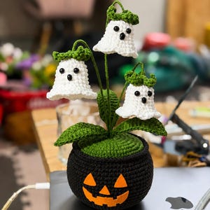 May include: A crocheted plant featuring three white ghost-shaped flowers with black bead eyes and green stems, leaves, and caps. The plant is potted in a round black crocheted base decorated with an orange felt jack-o'-lantern face design.