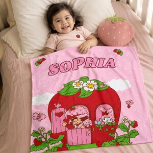 Puede incluir: Una manta de forro polar rosa suave con el nombre SOPHIA en letras negritas. El dise&ntilde;o incluye una gran casa con forma de fresa roja con hojas verdes y flores blancas, ocupada por un personaje y un conejo, rodeada de peque&ntilde;as fresas.