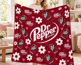 Puede incluir: Una manta de forro polar granate con un gran logotipo blanco de Dr. Pepper y el texto Est. 1885. La manta está decorada con un patrón repetitivo de flores blancas tipo margarita, pequeños corazones rosas e ilustraciones de latas de refresco.