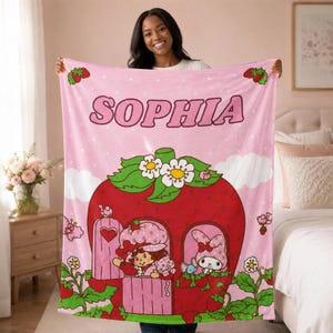Puede incluir: Una manta de forro polar rosa claro con el nombre SOPHIA en letras grandes en la parte superior. El dise&ntilde;o incluye una gran casa de fresa roja con hojas verdes y flores blancas, que contiene personajes de dibujos y detalles de fresas.