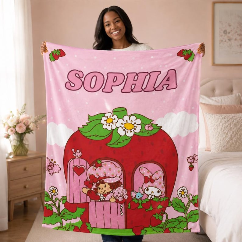 Puede incluir: Una manta de forro polar rosa claro con el nombre SOPHIA en letras grandes en la parte superior. El dise&ntilde;o incluye una gran casa de fresa roja con hojas verdes y flores blancas, que contiene personajes de dibujos y detalles de fresas.