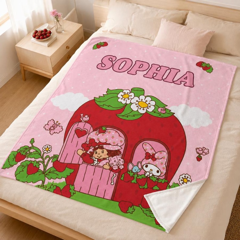 Puede incluir: Una manta de forro polar rosa claro con el nombre SOPHIA en letras grandes. El dise&ntilde;o incluye una casa grande con forma de fresa con hojas verdes y flores blancas, rodeada de plantas de fresa, un personaje de dibujos y un conejo.