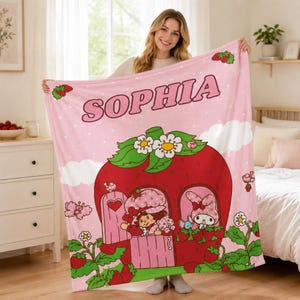 Puede incluir: Una manta de forro polar rosa suave con el nombre SOPHIA en letras grandes en la parte superior. El dise&ntilde;o incluye una casa de fresa roja grande con hojas verdes y flores blancas, con un personaje y un conejo visibles en las ventanas.