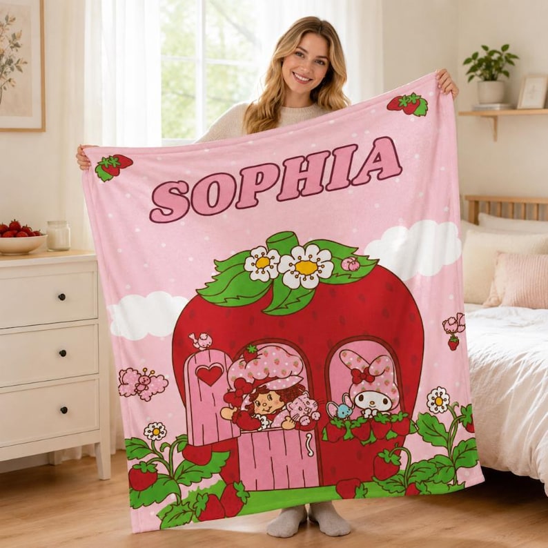 Puede incluir: Una manta de forro polar rosa suave con el nombre SOPHIA en letras grandes en la parte superior. El dise&ntilde;o incluye una casa de fresa roja grande con hojas verdes y flores blancas, con un personaje y un conejo visibles en las ventanas.