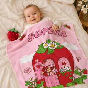 Puede incluir: Una manta de beb&eacute; de color rosa suave con el nombre SOPHIA en letras moradas, un gr&aacute;fico grande de una casa de fresa roja con personajes de Strawberry Shortcake y My Melody, flores blancas, nubes y un peque&ntilde;o juguete de peluche de fresa.
