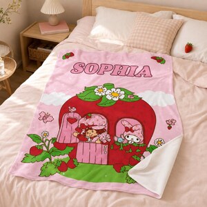 Puede incluir: Una manta de forro polar rosa suave con el nombre SOPHIA en letras negritas en la parte superior. El dise&ntilde;o incluye una casa grande con forma de fresa roja con hojas verdes, flores blancas, un personaje, un conejo y motivos de fresas.