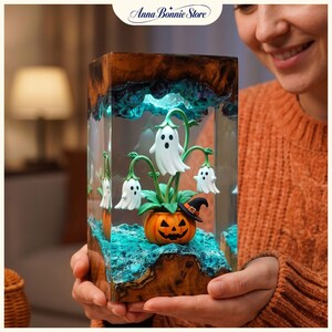 May include: A rectangular resin lamp featuring a carved wooden top and base with a glowing teal interior. Inside, a miniature orange jack-o'-lantern planter holds green stems with three white ghost-shaped flowers and a small black witch hat.