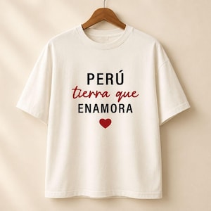 Puede incluir: Una camiseta de manga corta color blanco hueso con el texto negro PERÚ, el texto en cursiva roja tierra que, el texto negro ENAMORA y un pequeño icono de corazón rojo centrado en el pecho. La camiseta se muestra en una percha de madera.