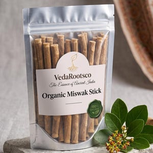Op de afbeelding: Een zilveren stazak gevuld met lichtbruine biologische Miswak-stokjes. Het etiket bevat de tekst VedaRootsco, The Essence of Ancient India, Organic Miswak Stick en een groen 100% Organic-zegel. Een klein groen takje met blaadjes ligt ernaast.