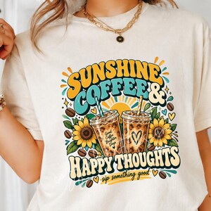 Może przedstawiać: Kremowy t-shirt z nadrukiem graficznym z napisem SUNSHINE COFFEE & HAPPY THOUGHTS. Wzór zawiera dwa kubki mrożonej kawy, słoneczniki, ziarna kawy i ikonę słońca. Na jednym z kubków znajduje się napis Good Coffee Brighter Days.