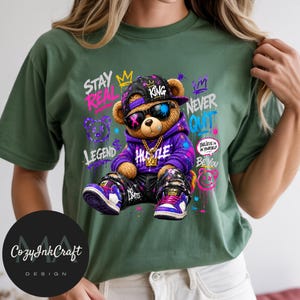 Może przedstawiać: Oliwkowy t-shirt z grafiką brązowego pluszowego misia w fioletowej bluzie z kapturem, czarnej czapce, okularach przeciwsłonecznych i złotym łańcuchu. Projekt zawiera napisy w stylu graffiti: STAY REAL, KING, NEVER QUIT, LEGEND, BE YOU oraz NO LIMITS.