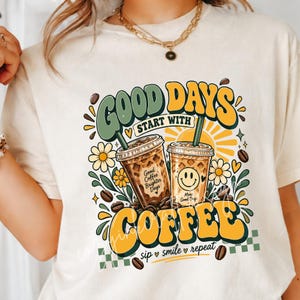 Może przedstawiać: Kremowy t-shirt z grafiką przedstawiającą dwa kubki mrożonej kawy, żółte stokrotki i ziarna kawy. Napis głosi "GOOD DAYS START WITH COFFEE" oraz "sip smile repeat". Projekt zawiera żółte słońce i zielone akcenty dekoracyjne.