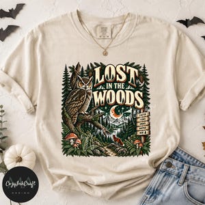 Może przedstawiać: Kremowy t-shirt z nadrukiem sowy siedzącej na gałęzi w lesie. Napis głosi LOST IN THE WOODS, a w grafice widać półksiężyc, małego lisa, grzyby oraz drewniany drogowskaz z wyrazami QUIETER, HIGHER, KINDER, HAPPIER.