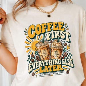 May include: A cream-colored t-shirt featuring a graphic print of two iced coffee cups, a sunburst, and coffee beans. The text reads COFFEE FIRST EVERYTHING ELSE LATER, GOOD COFFEE BRIGHTER DAYS, Coffee Makes Everything Better, and SLOW MORNINGS STRONG COFFEE.