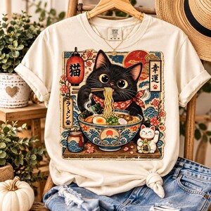 Może przedstawiać: Kremowy t-shirt z grafiką przedstawiającą czarnego kota jedzącego ramen z miski w falisty wzór. Projekt zawiera czerwoną latarnię, kwiaty wiśni, kota szczęścia oraz japoński tekst. Koszulka ma krótkie rękawy i okrągły dekolt.