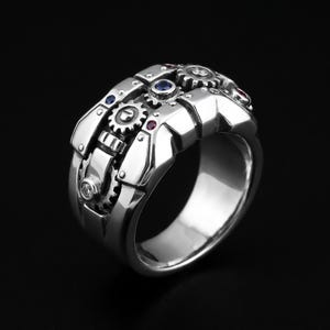 Puede incluir: Un anillo de tono plateado con un diseño de engranajes mecánicos y piedras circulares incrustadas en azul, rojo y transparente. La banda tiene una construcción ancha y segmentada con detalles metálicos en relieve que imitan componentes de relojería.