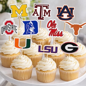 May include: Cupcakes with white frosting and decorative toppers featuring college team logos. Logos include "M", "A&M", "A", "Ohio State", "Duke", "Ole Miss", "Texas", "Miami", "LSU", and "Georgia". The cupcakes are arranged on a white cake stand.