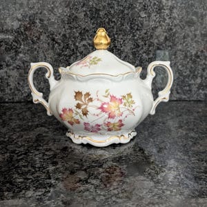 May include: A white porcelain sugar bowl with a lid, featuring a delicate pattern of autumn-colored maple leaves in shades of brown, pink, and green. The bowl has two ornate handles, a gold-toned finial on the lid, and gold trim on the edges.