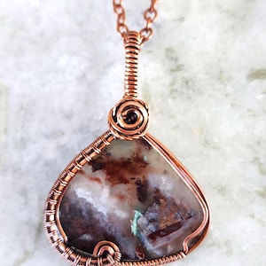 May include: A copper wire-wrapped pendant necklace featuring a teardrop-shaped stone. The stone has a mix of brown, white, and green hues. The necklace chain and wire wrapping are a warm copper color, creating a handcrafted look.