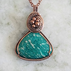 May include: A handcrafted copper pendant necklace featuring a teardrop-shaped, teal-colored stone with white veining. The stone is encased in copper wire, with a decorative top section and a copper chain.