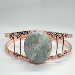 May include: A rose gold-toned cuff bracelet featuring an oval-shaped, mottled grey and green stone centerpiece. The metal band displays intricate filigree patterns and decorative wire-wrapped scrollwork accents on both sides of the central stone.
