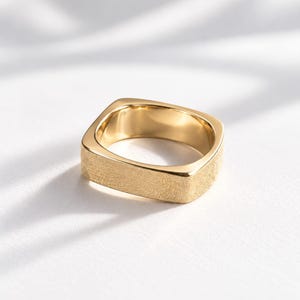 Könnte beinhalten: Ein goldener Ring mit quadratischem Band, das eine texturierte, matte Außenfläche und eine glatte, polierte Innenfläche aufweist. Der Ring hat abgerundete Ecken und ein breites, flaches Profil in einer metallisch gelben Farbe.