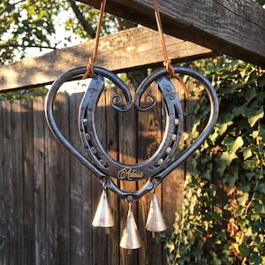 May include: A heart-shaped wall hanging crafted from two dark grey metal horseshoes, featuring a tan leather cord for suspension. The piece includes three small, conical metallic bells hanging from the bottom and the name Adessa in gold script.