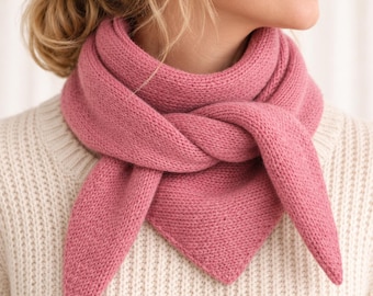 May include: A solid dusty rose pink triangular knit scarf with a textured garter stitch pattern. The scarf is wrapped around the neck and tied in a knot at the front, with two pointed ends hanging down over a cream-coloured ribbed knit jumper.