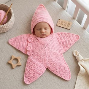 Puede incluir: Un capullo de ganchillo para bebé en color rosa claro con forma de estrella de cinco puntas y capucha a juego. La prenda cuenta con dos botones circulares en un brazo y se acompaña de un mordedor de madera y una etiqueta de madera con el texto COZY STAR COCOON.