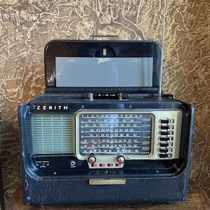May include: A black Zenith Trans-Oceanic portable radio featuring a rectangular dial with frequency markings, a vertical antenna, a top-mounted handle, and a flip-up lid displaying Zenith log charts. The front panel includes a speaker grille and buttons.