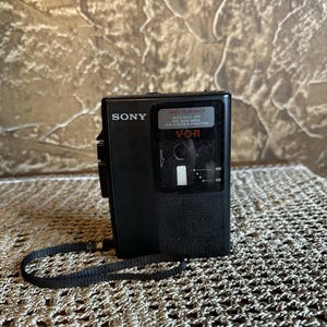 May include: A black Sony portable cassette recorder featuring a VOR system, fast playback, and a microphone sensitivity mode. The device includes a black wrist strap and a clear window displaying internal mechanics with numerical markings 50 and 100.