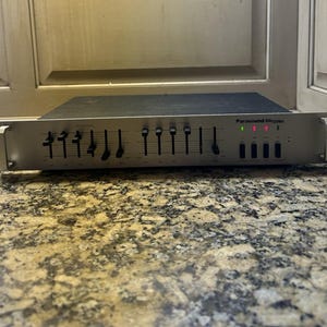Könnte beinhalten: Ein silberner Parasound EQ1200b Audio-Equalizer mit einer Reihe schwarzer vertikaler Schieberegler zur Frequenzanpassung, vier schwarzen Drucktasten und drei kleinen leuchtenden Anzeigeleuchten auf der Vorderseite mit Rack-Griffen.