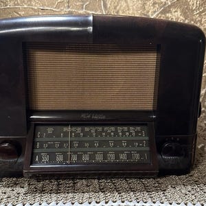 May include: A vintage RCA Victor tabletop radio featuring a dark brown, polished bakelite casing with a rectangular speaker grille covered in tan fabric. The front panel displays a glass-covered tuning dial with three frequency bands and knobs.