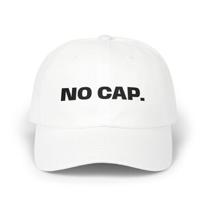May include: A white baseball cap featuring the text NO CAP. printed in bold black capital letters across the front panel. The hat has a structured crown, a curved brim, and a button at the top centre, maintaining a clean and simple design.